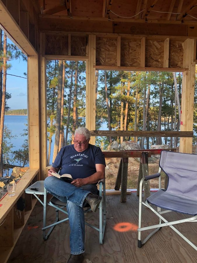 Ron reading in a camp chair in the framed-in porch, lake behind